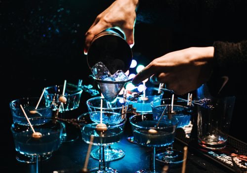 Close-up of a bartender's hands pouring cocktails through a strainer over ice in a dimly lit bar, capturing the essence of nightlife and mixology in a lively party atmosphere.